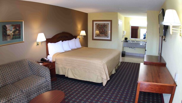 A hotel room with a large bed, a sofa, a small table, a desk, a mirror, and a bathroom with a sink and toilet. - Loyalty Inn Hawkinsville (Hawkinsville)