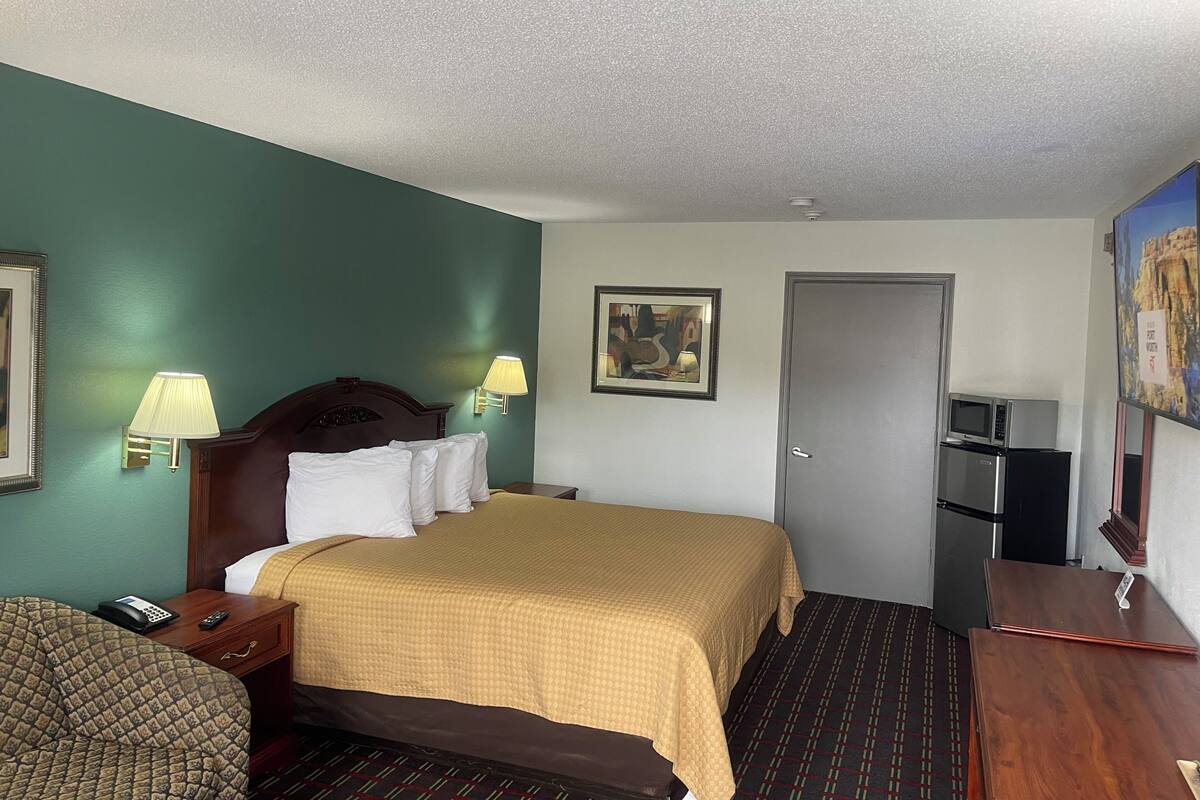 A hotel room with a bed, bedside lamps, a desk, a chair, a door, a refrigerator, and a microwave.
