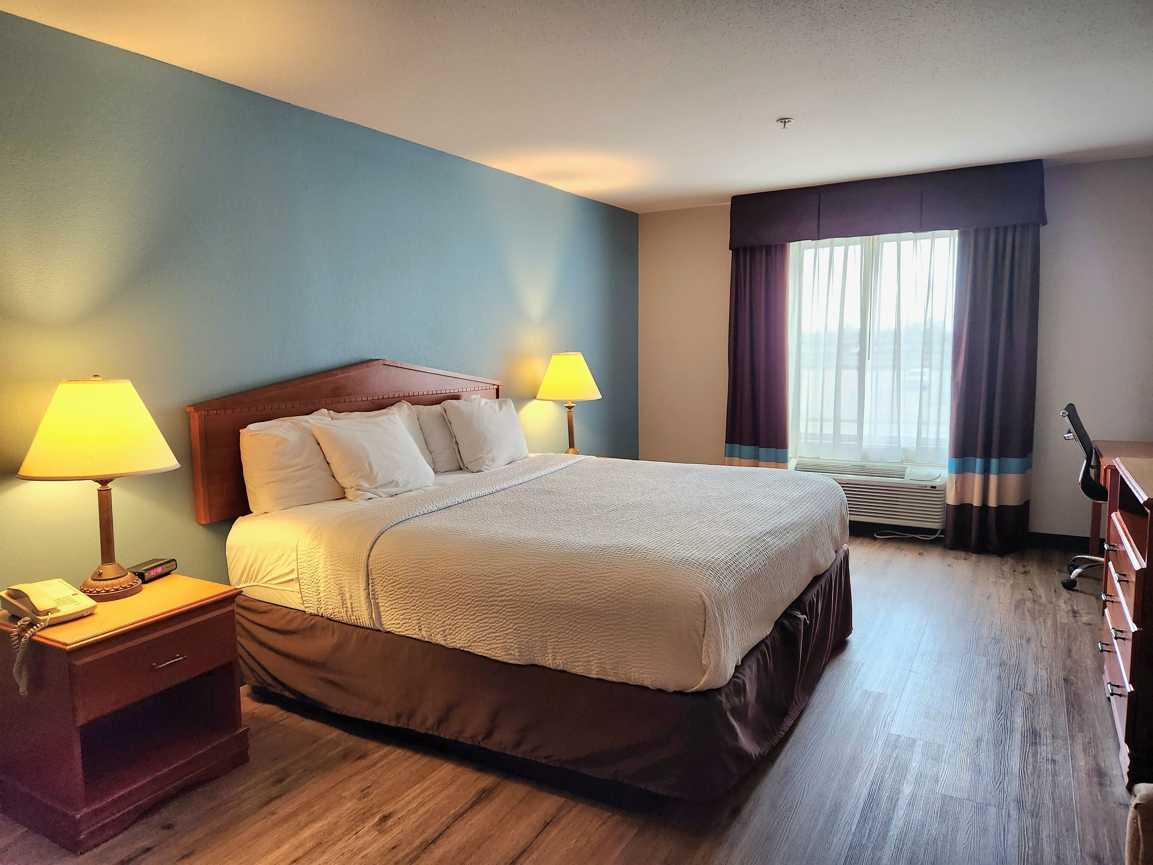Habitación de hotel con cama, lámparas de noche, mesita de noche, escritorio y ventana con cortinas.