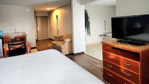 A hotel room with a bed, a TV on a wooden dresser, a desk with a chair, a sofa, and a plant.
