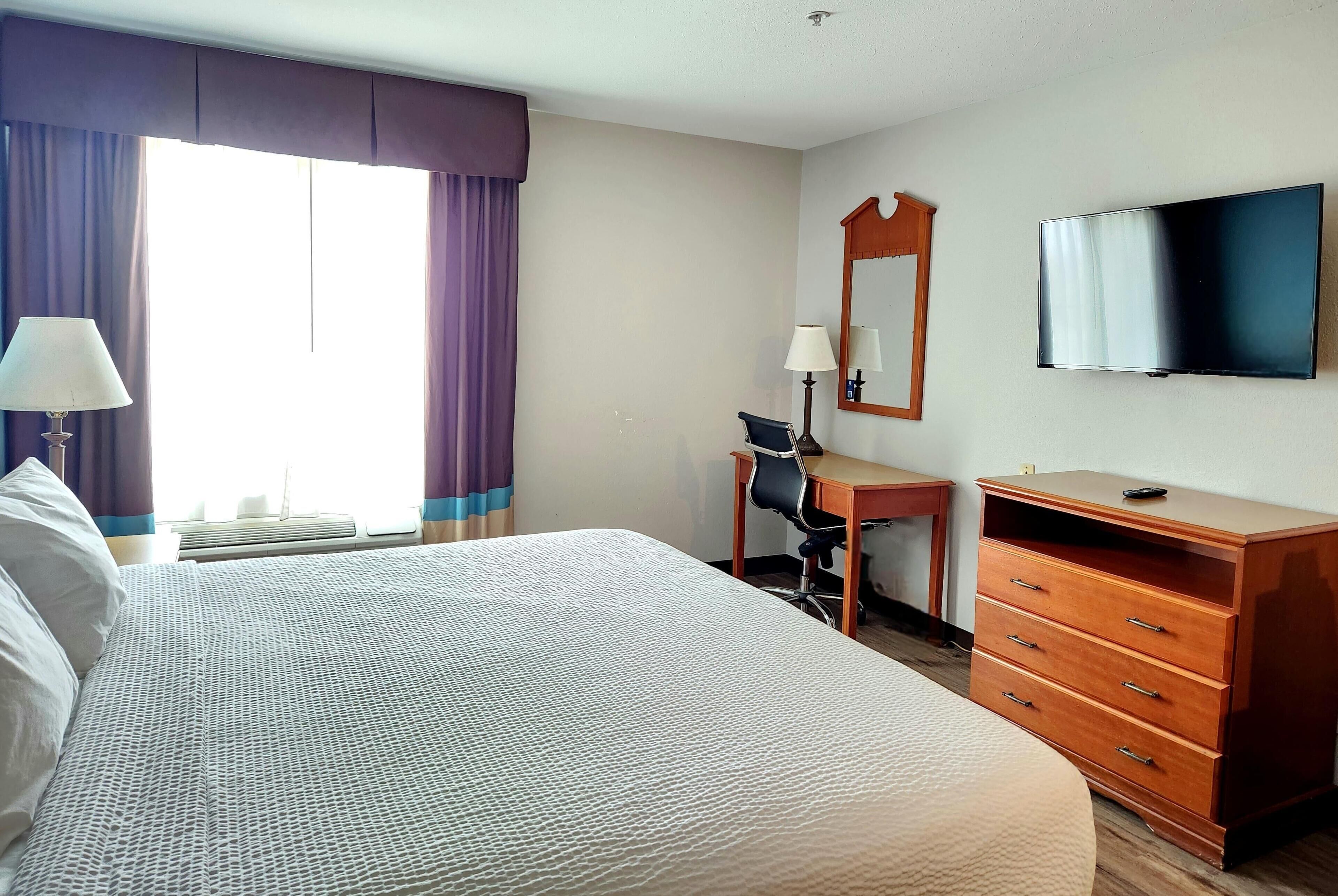 A hotel room with a large bed, a desk with a chair, a dresser, a television, and a window with curtains.