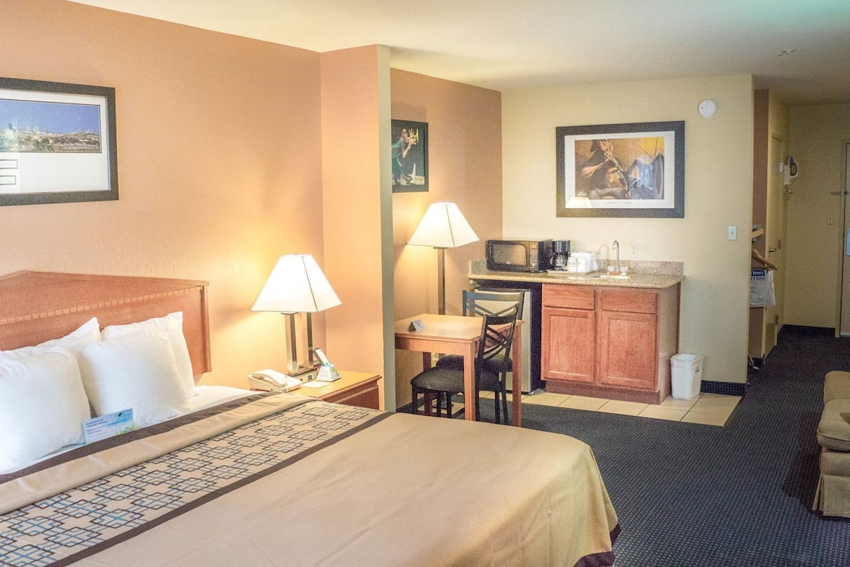A hotel room with a bed, a desk with a lamp, a chair, a small kitchenette, and a door leading to another room.