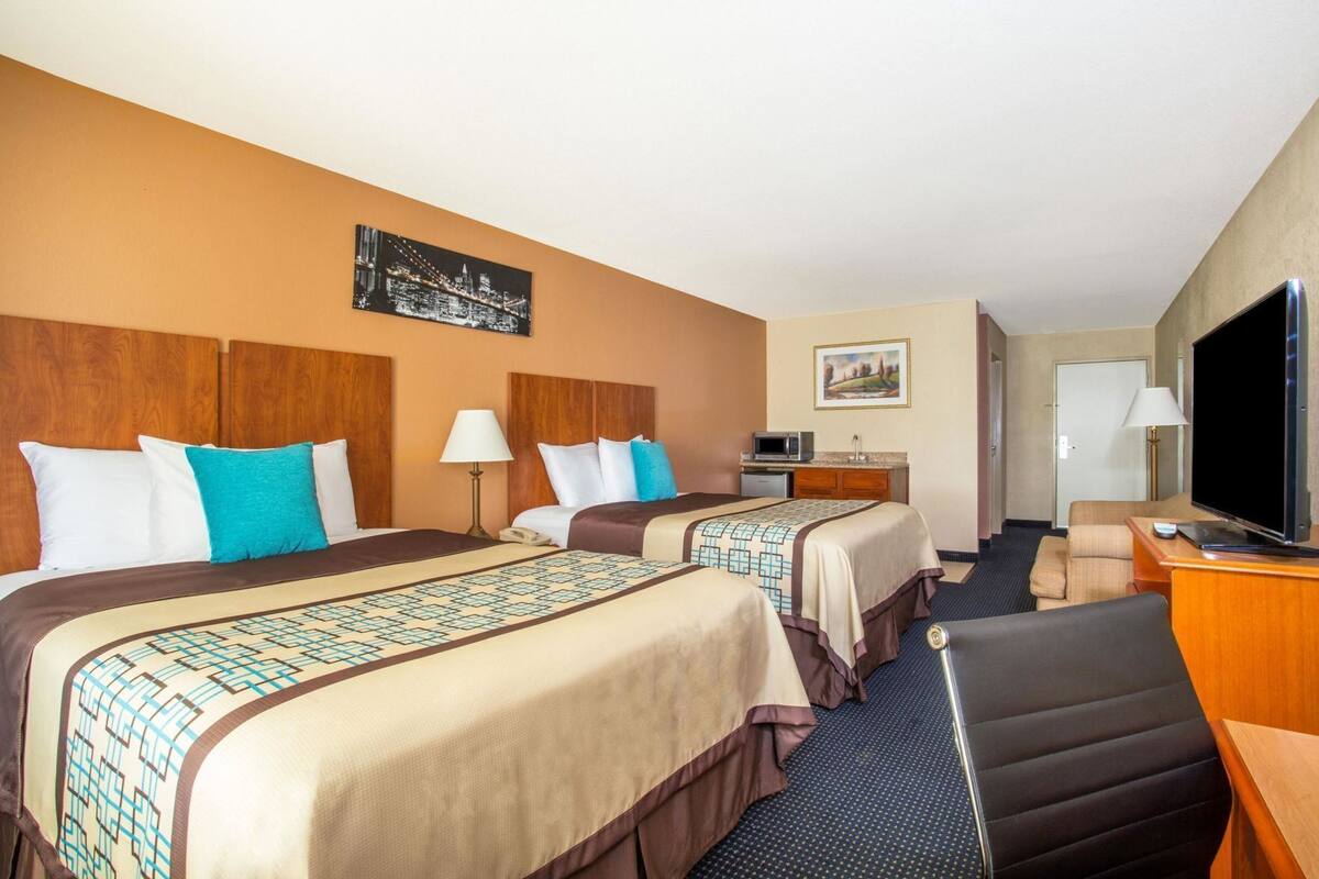 A hotel room with two beds, a desk, a television, and a small kitchenette.