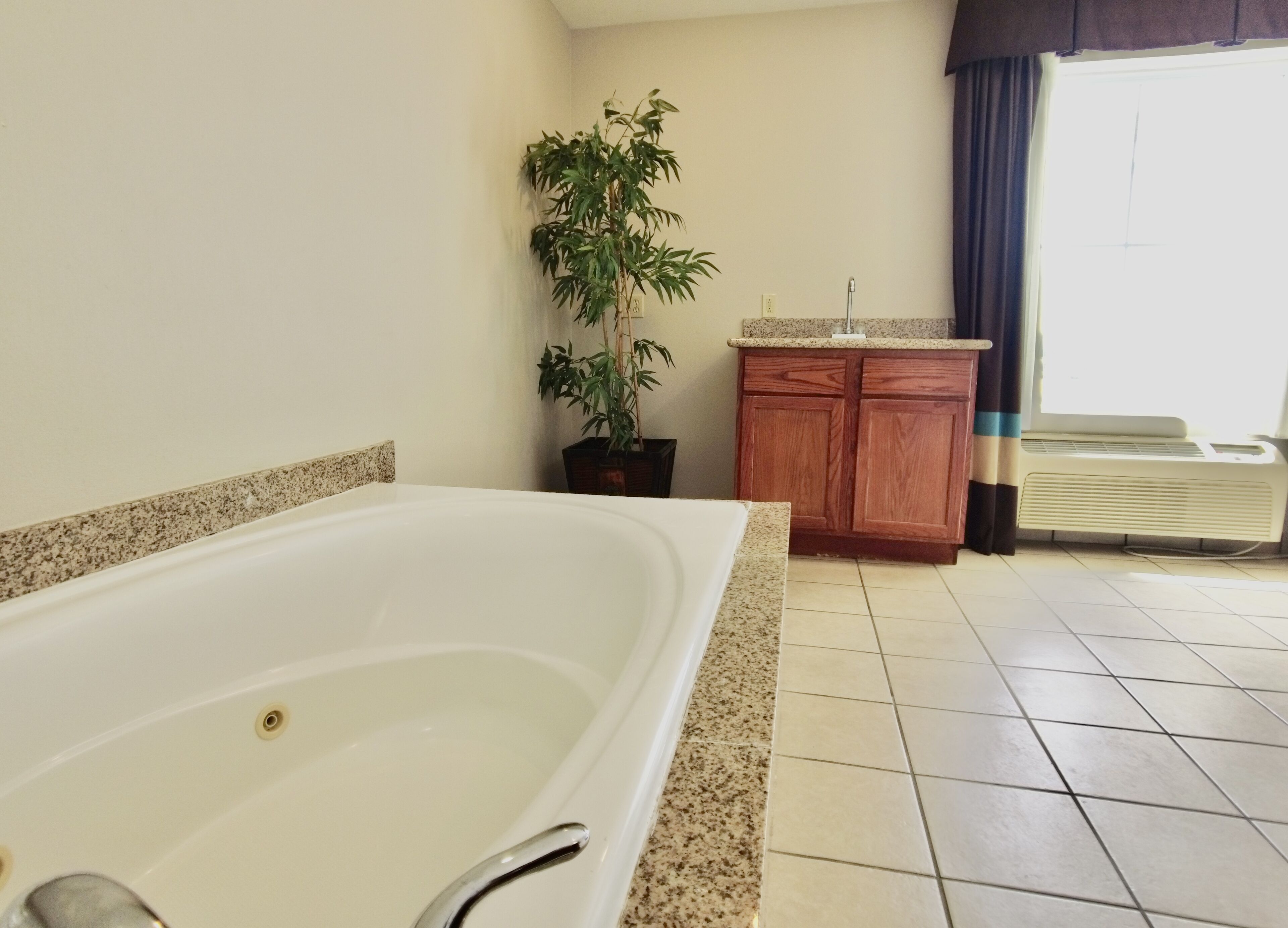 A bathroom with a large bathtub, a wooden cabinet, a potted plant, and a window with curtains.
