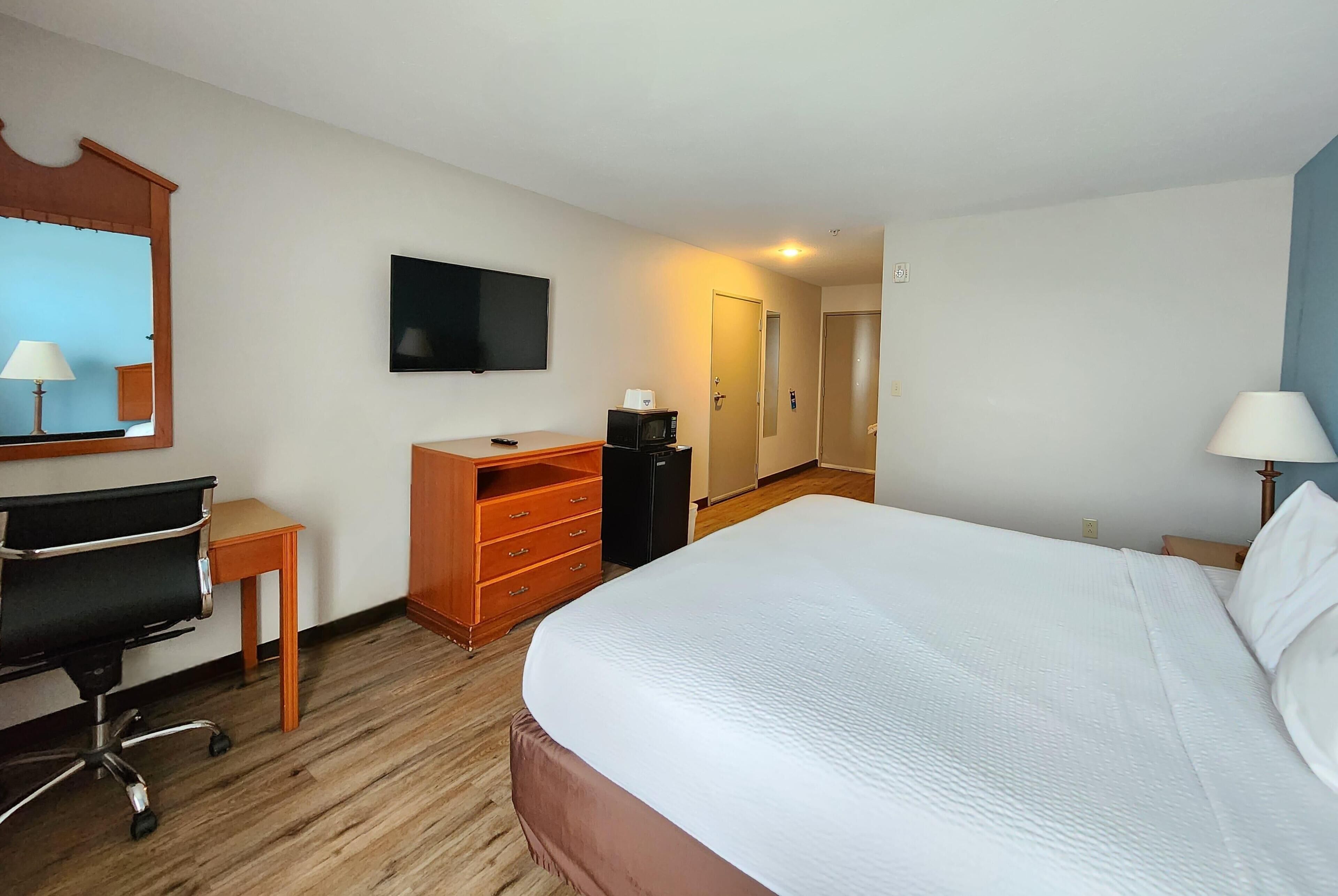 A hotel room with a large bed, a desk with a chair, a television, a dresser, and a door leading to another room.