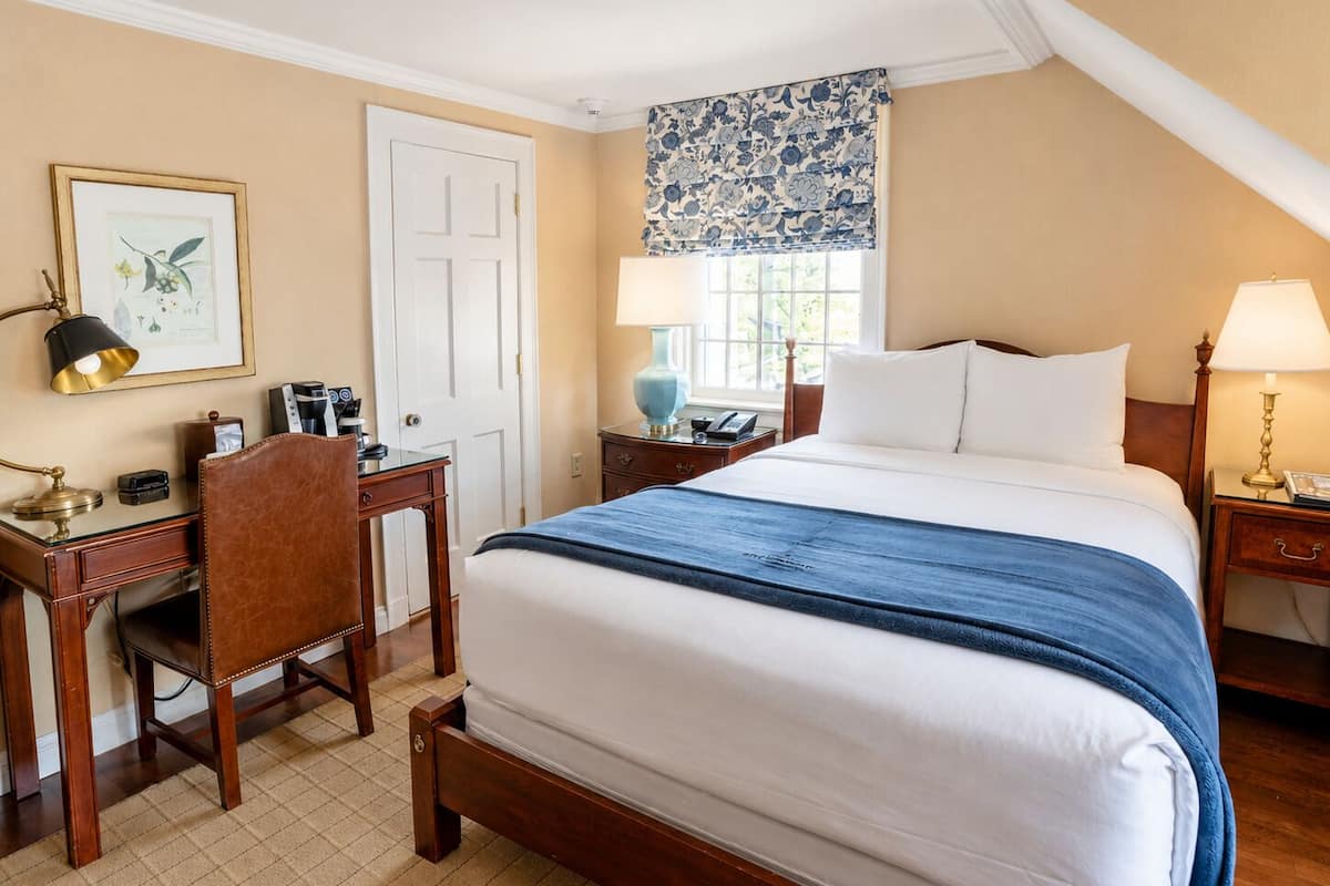 Une chambre d’hôtel équipée d’un lit, d’un bureau, d’une chaise, de tables de chevet, d’une lampe et d’une fenêtre donnant sur un paysage agréable.