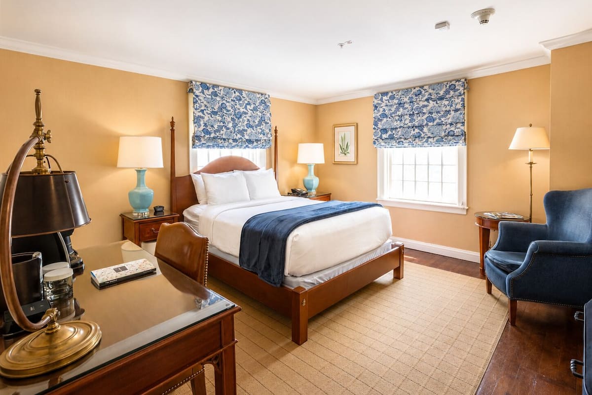 Une chambre d’hôtel avec un lit en bois, un bureau avec une lampe, une chaise et une fenêtre avec des rideaux à motifs.