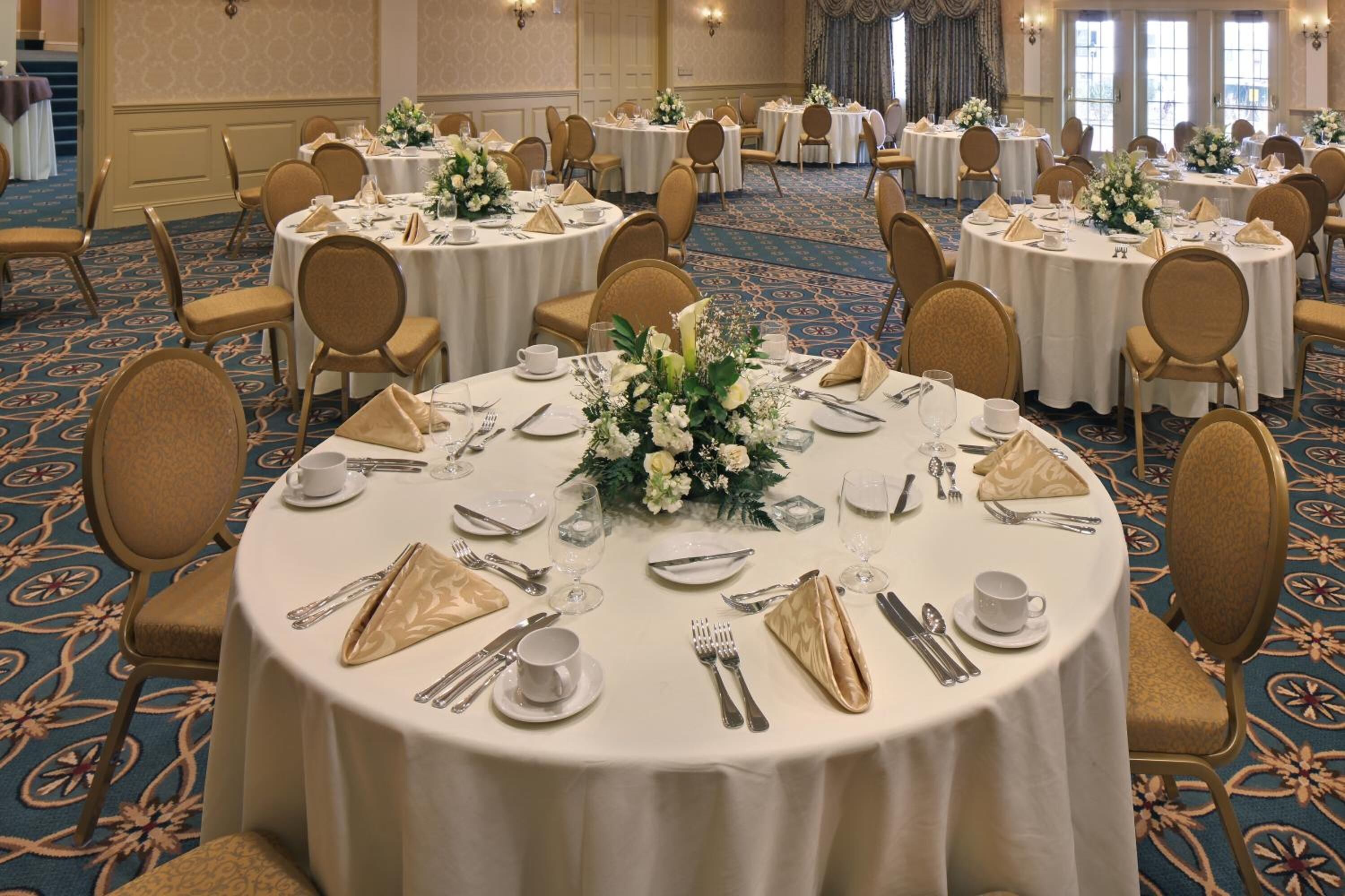 A banquet hall with round tables set for dining, each with white tablecloths, gold napkins, and floral centerpieces.