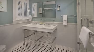 A bathroom with a double sink, a glass shelf, a towel rack, and a shower area.