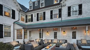 A spacious outdoor seating area with a fire feature, white brick walls, and multiple windows with shutters. - Inn on Boltwood (Amherst)