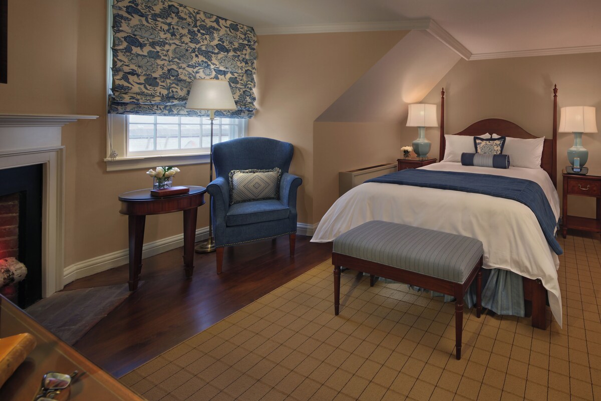 A bedroom with a bed, a chair, a nightstand, a lamp, and a fireplace.