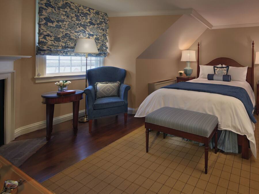 A bedroom with a bed, a chair, a nightstand, a lamp, and a fireplace.