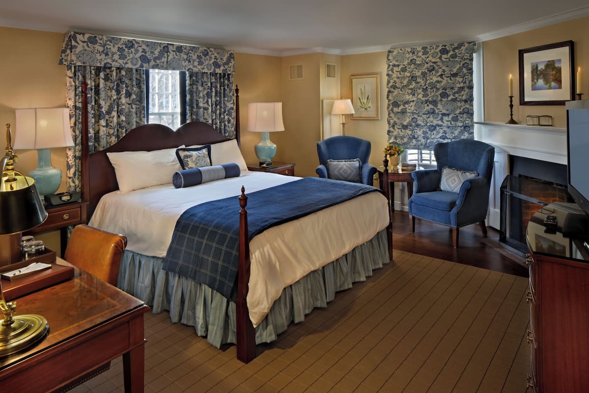 Une chambre d’hôtel avec un grand lit, deux fauteuils, un bureau, une cheminée et une fenêtre avec des rideaux.