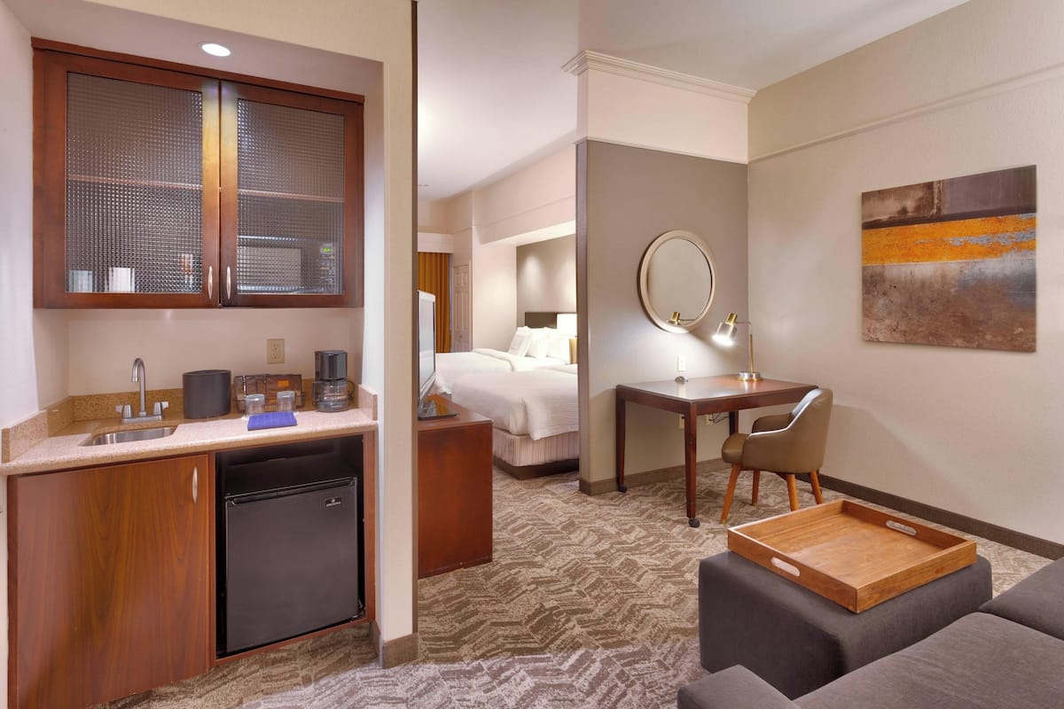 A hotel room with a kitchenette, a bed, a desk with a chair, a mirror, and a painting on the wall.