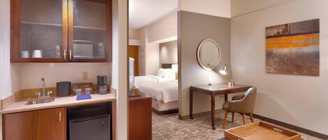 A hotel room with a kitchenette, a bed, a desk with a chair, a mirror, and a painting on the wall.