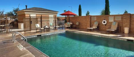 A clear pool with a lifeguard chair and a red umbrella, surrounded by lounge chairs and a brick wall.