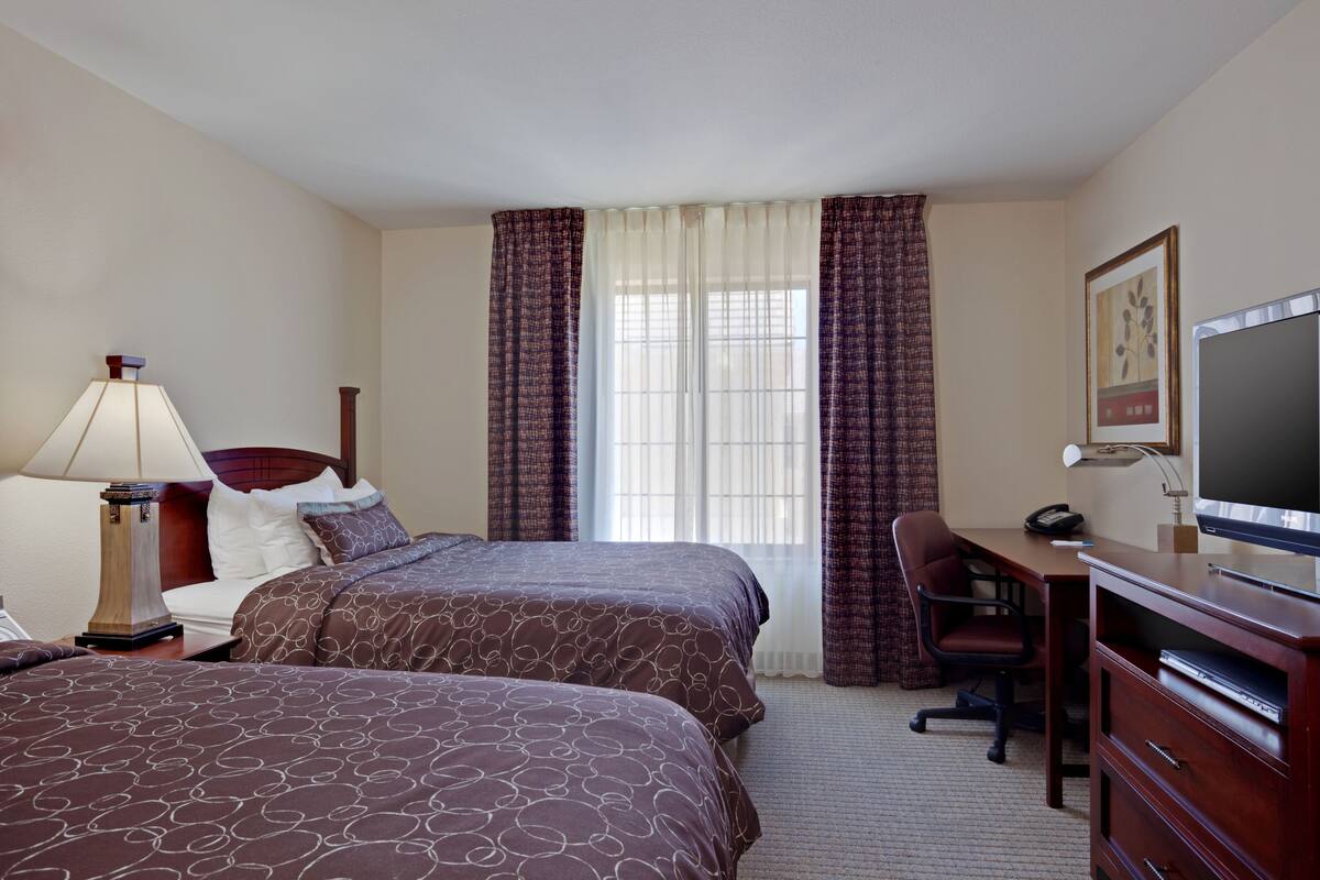A hotel room with two beds, a desk, a chair, a TV, and a lamp.