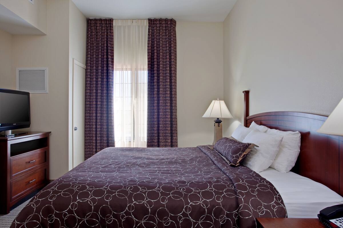 A hotel room with a large bed, a TV on a wooden dresser, a lamp, and a window with curtains.