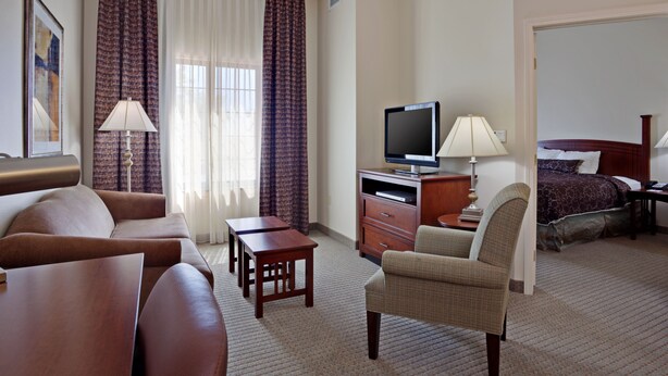A hotel room with a sofa, armchair, TV, desk, and a bed with a headboard. - Staybridge Suites Palmdale by IHG (Palmdale)