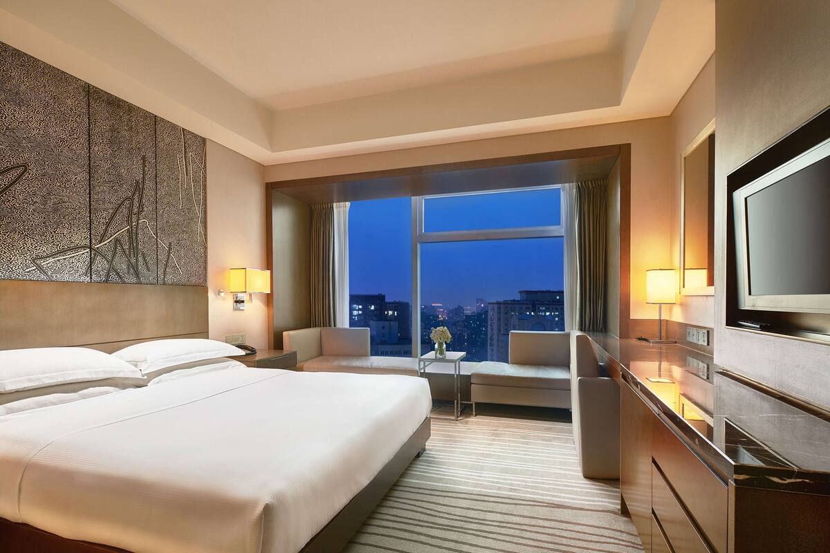 A hotel room with two beds, a desk, a chair, a TV, and a large window with a city view.