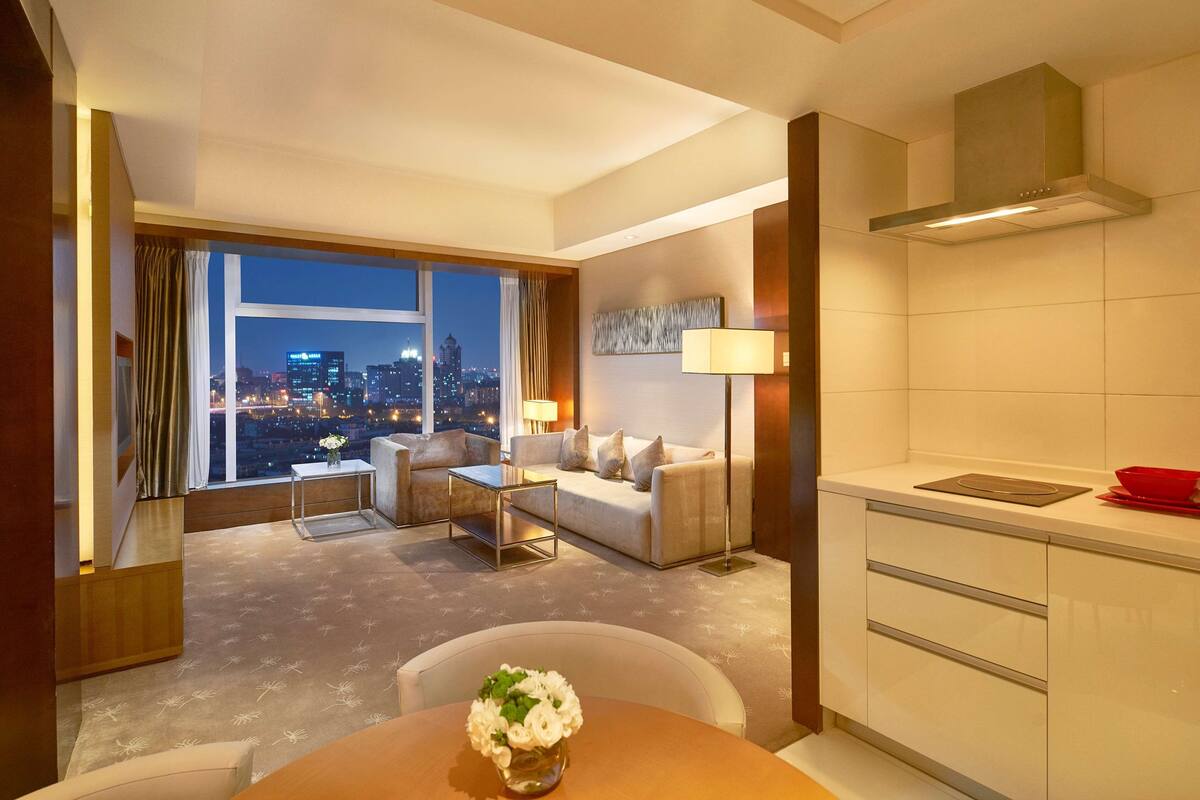 A hotel room with a city view, a sofa, a dining table with a vase of flowers, a lamp, and a kitchenette.