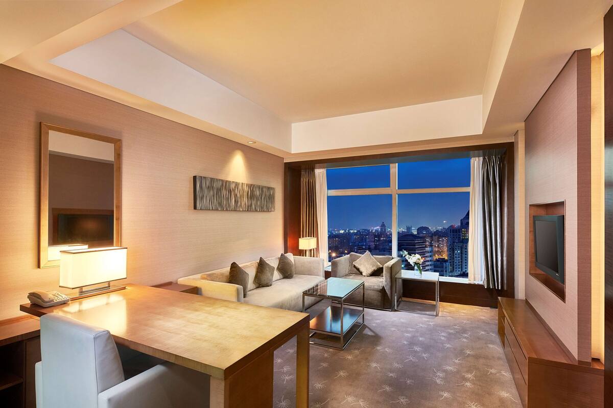 A hotel room with a sofa, two armchairs, a coffee table, a TV, a desk with a phone, and a large window with a city view.