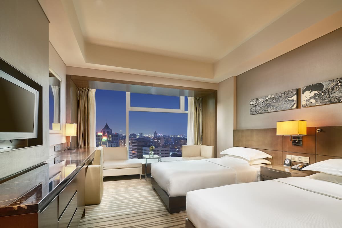 A hotel room with two beds, a TV, a desk, and a view of the cityscape.