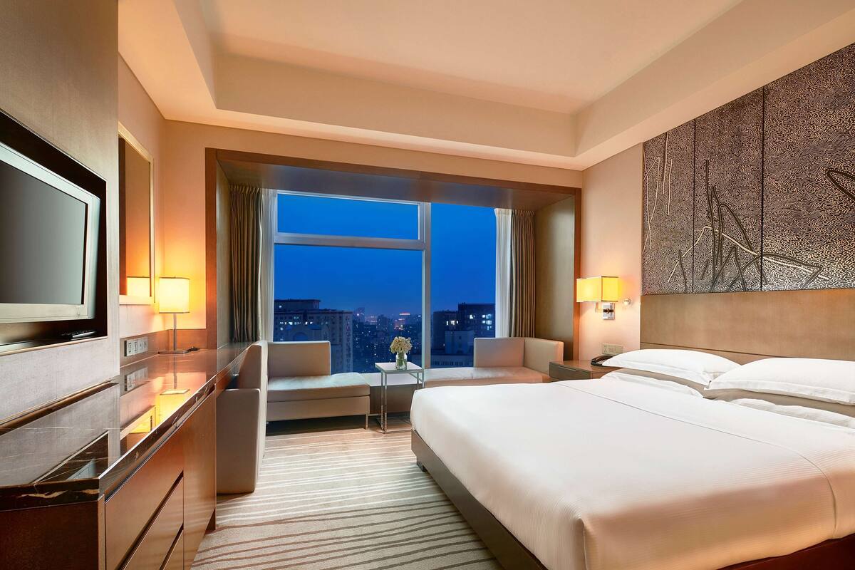 A hotel room with a large bed, a desk, a chair, a TV, and a window with a city view.