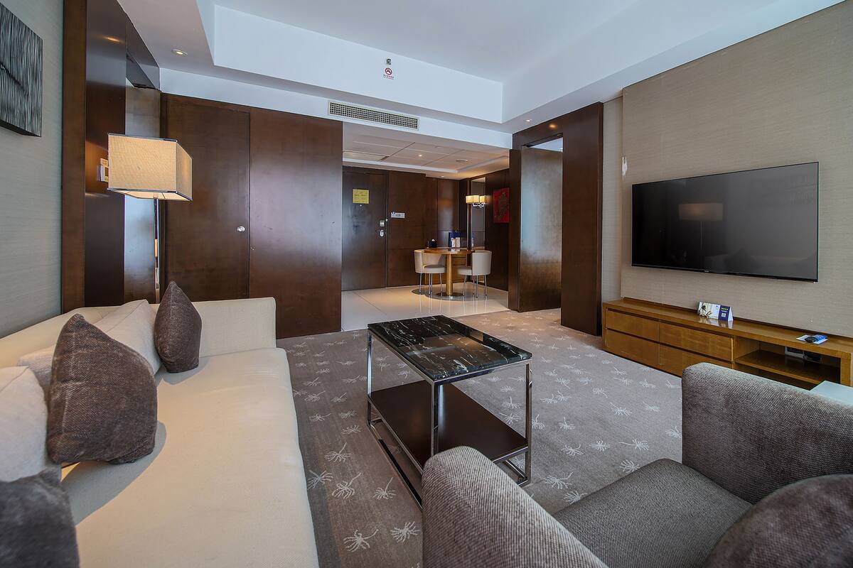 A modern hotel lobby with a sofa, armchairs, a coffee table, a TV, and a small dining area.