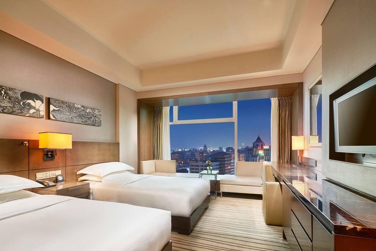 A hotel room with two beds, a desk, a TV, and a view of the cityscape at night.