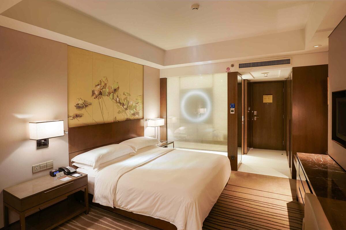 A hotel room with a bed, bedside lamps, a desk with a phone, a painting on the wall, a door with a sign, and a TV on the right.