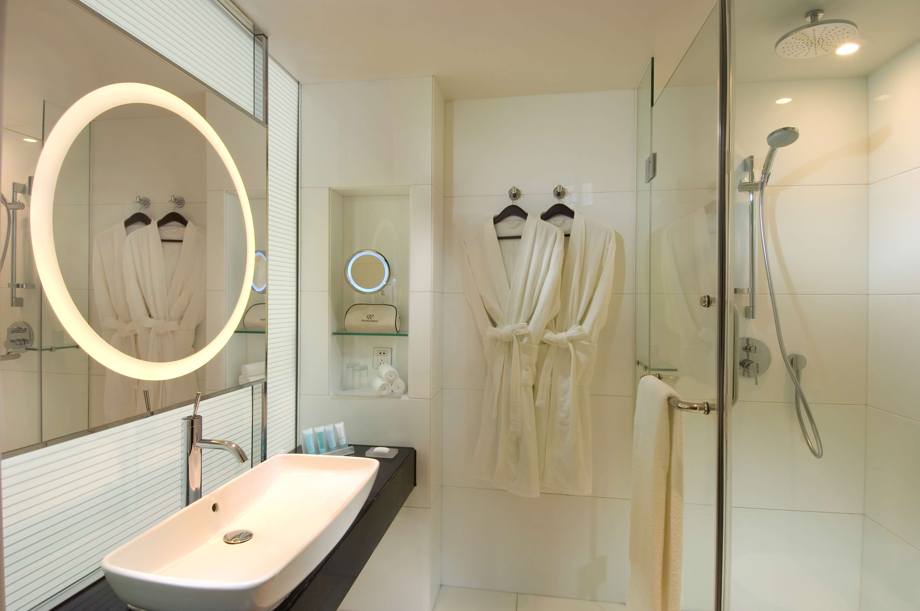 Une salle de bain moderne avec un grand miroir, un lavabo, une douche et deux peignoirs suspendus à des crochets.