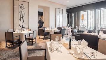 Un restaurante con mesas dispuestas para comer, un cuadro abstracto de gran tamaño en la pared y vistas de la ciudad a través de amplias ventanas.