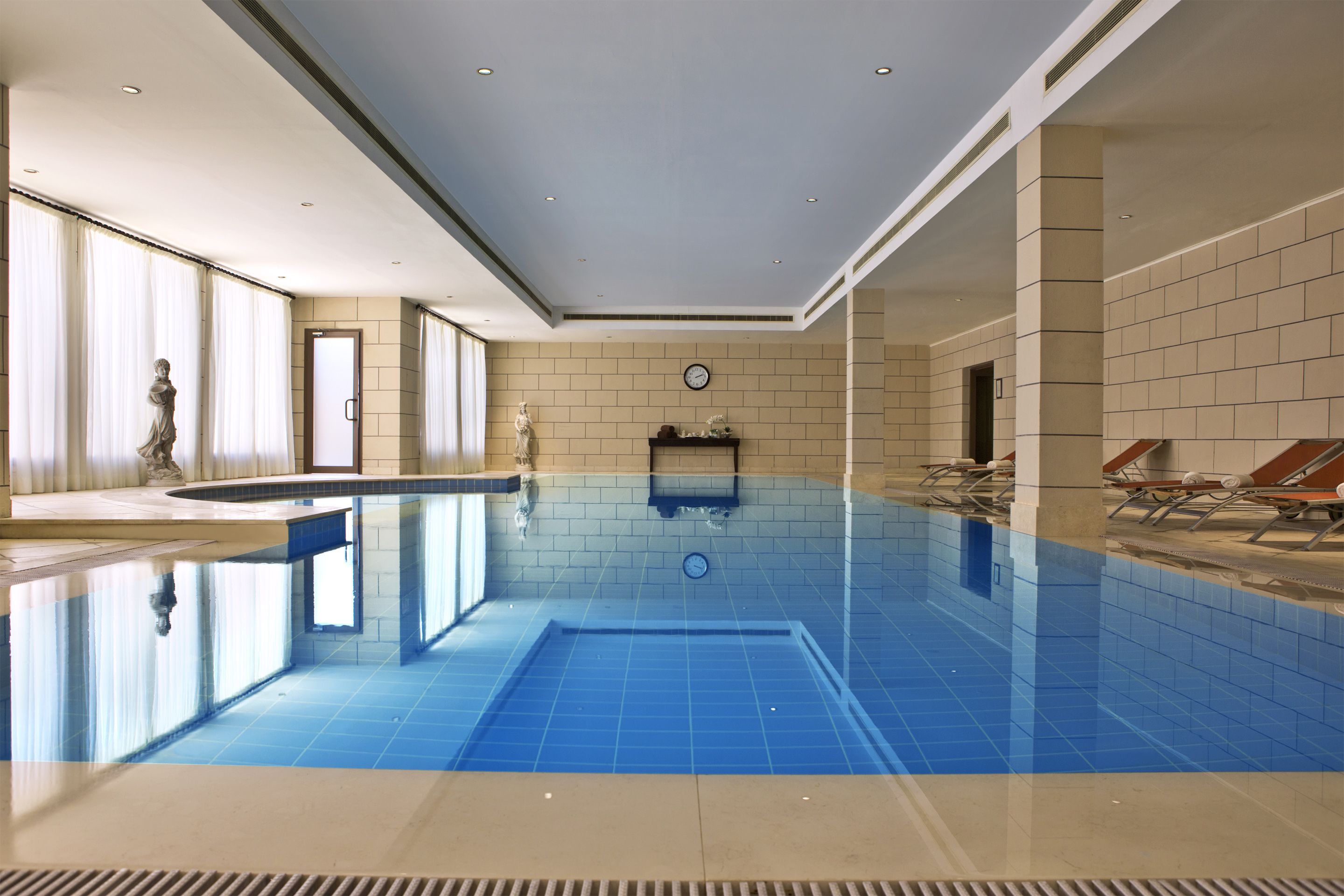A large indoor swimming pool with clear blue tiles, a statue near the entrance, and a clock on the wall.