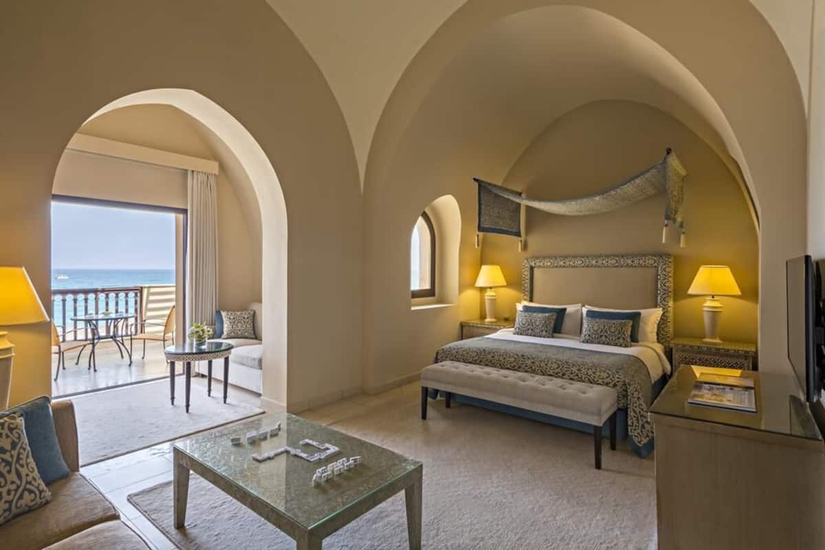 A hotel room with a large bed, a balcony with a view of the sea, a sitting area with a table and chairs, and a TV on a stand.