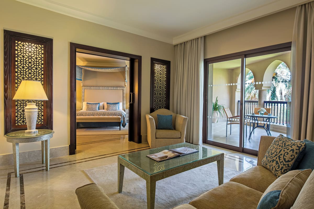 A hotel room with a bed, a sitting area with a sofa and armchair, a glass coffee table, and a view of the outdoor patio.