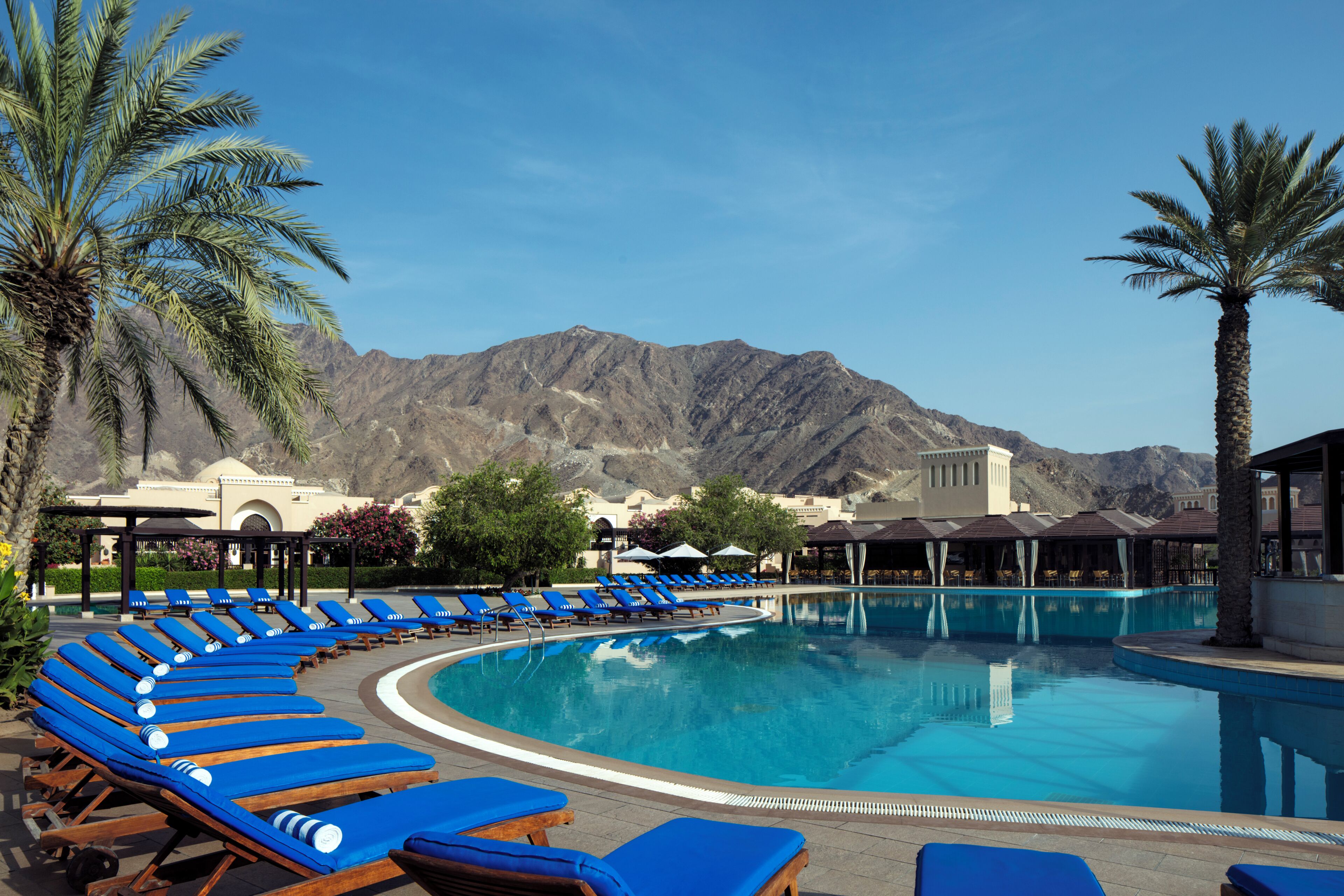 Una piscina con sillas reclinables azules, palmeras y montañas al fondo.