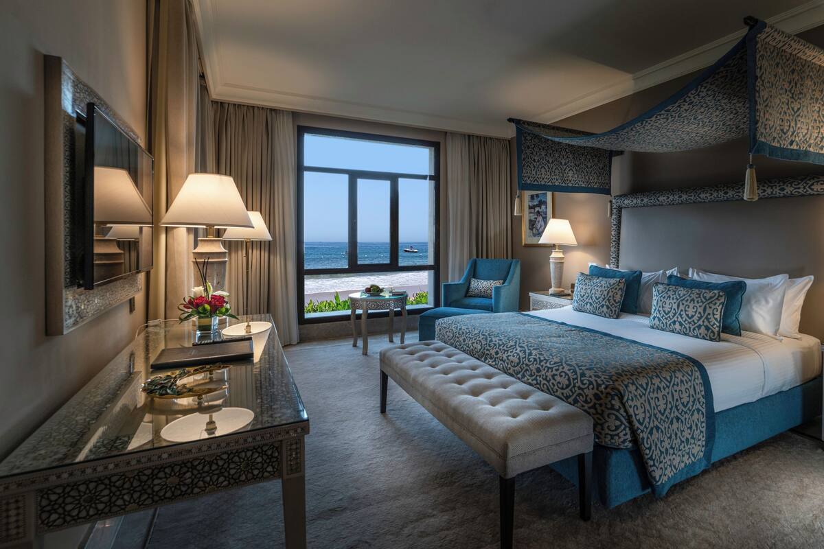 A hotel room with a large bed, two bedside tables, a bench, a TV, and a view of the sea.