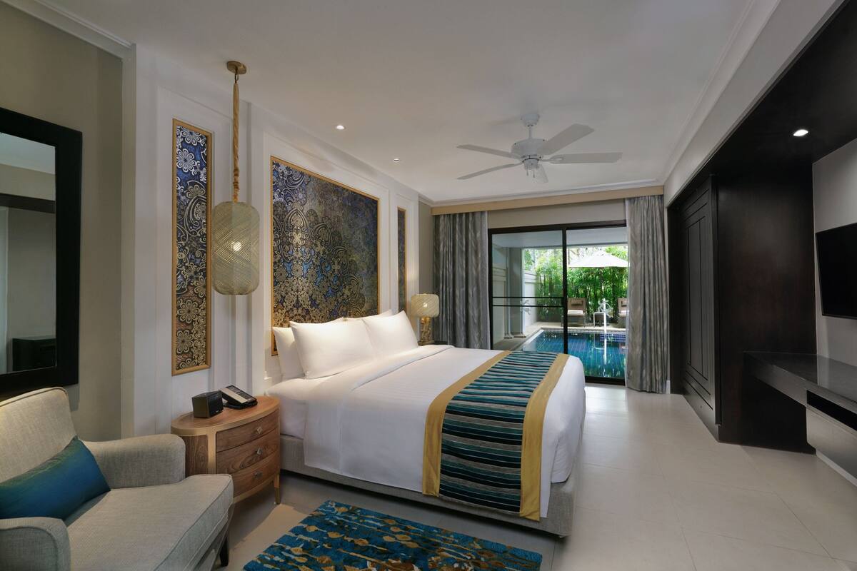 A hotel room with a large bed, a bedside table, a chair, a TV, a ceiling fan, and a view of a pool.