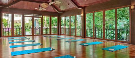 A spacious yoga studio with wooden floors and large windows offering a view of lush greenery.
