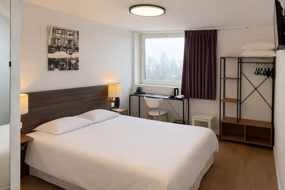 A hotel room with a large bed, a desk, a chair, a wardrobe, and a window with curtains.
