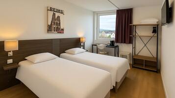 Ein Hotelzimmer mit zwei Betten, einem Schreibtisch, einem Stuhl, einem Fernseher, einem Regal und einem Fenster mit Blick auf Gebäude.