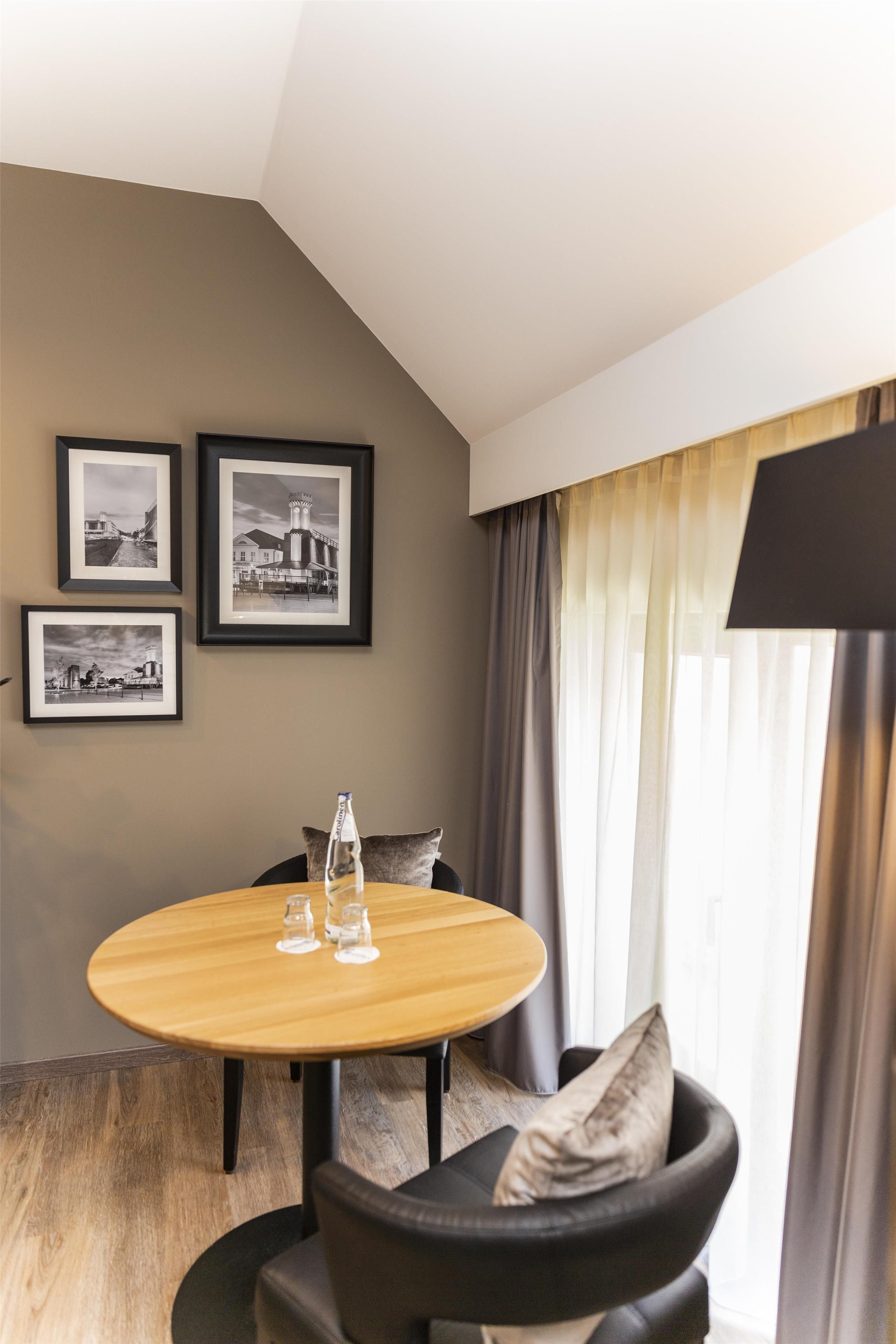 Une chambre d’hôtel avec une table ronde en bois, deux chaises, une lampe noire et trois photographies en noir et blanc encadrées accrochées au mur.