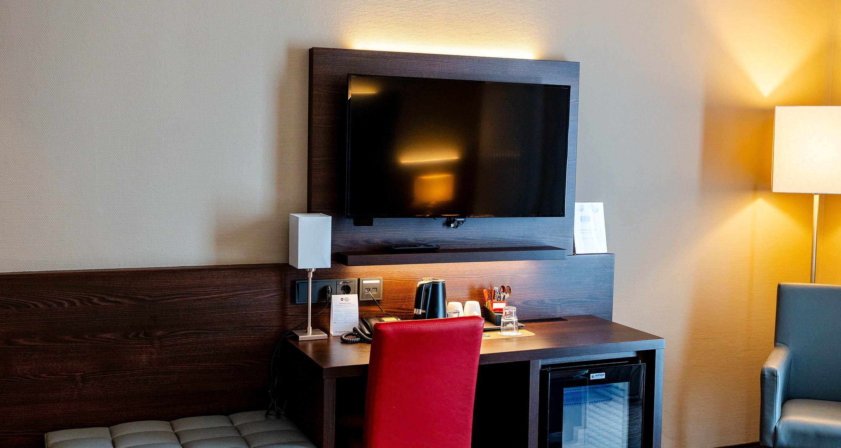 A hotel room with a flat-screen TV mounted on the wall, a desk with a telephone, a red chair, a minibar, and a small refrigerator.
