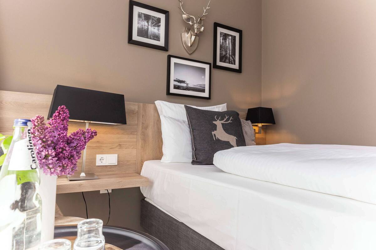 Une chambre d’hôtel avec une tête de lit en bois, un téléviseur à écran plat, un lit avec une literie blanche, un coussin décoratif à l’effigie d’un cerf et une tête de cerf accrochée au mur.