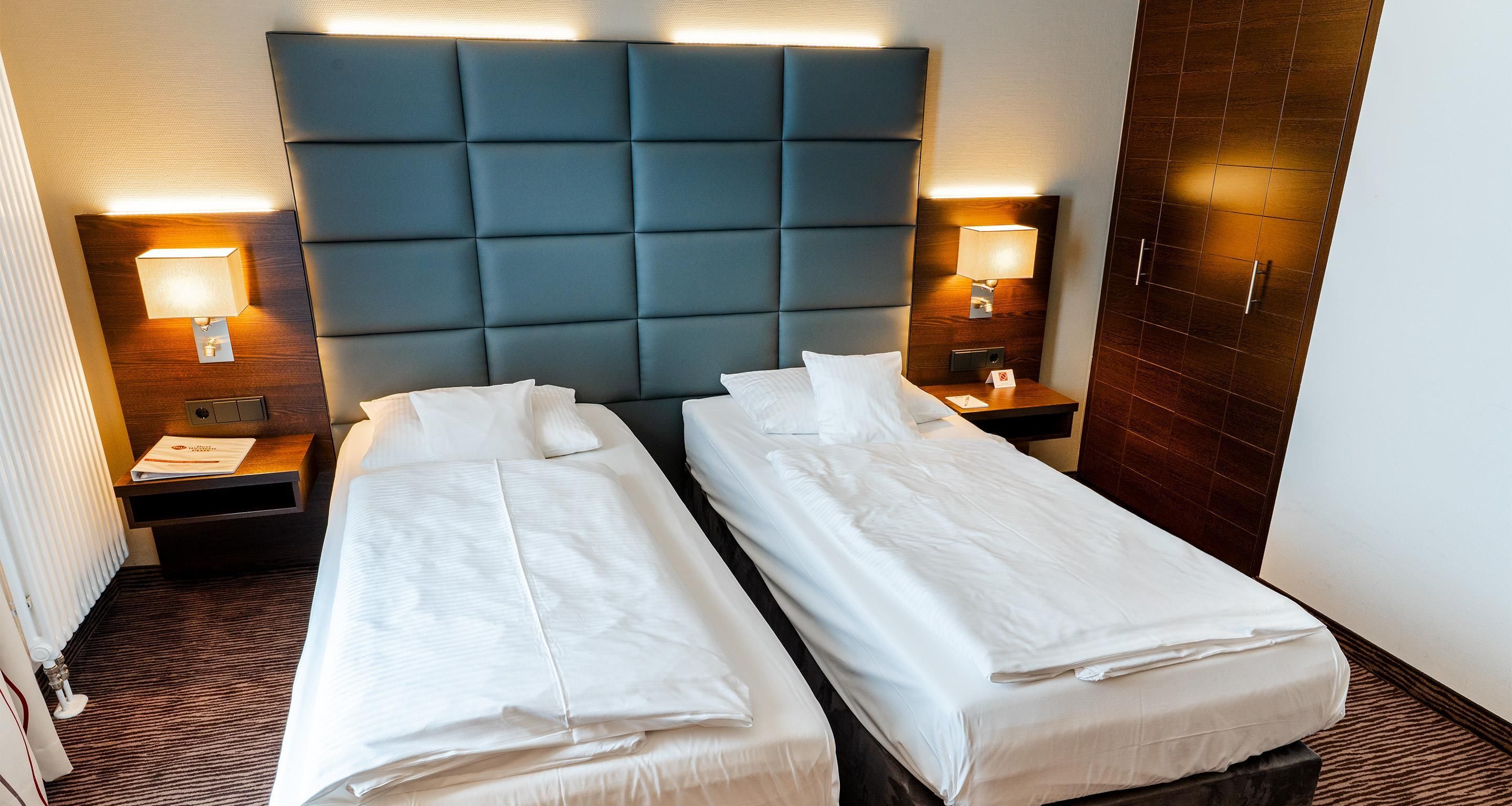 Ein Hotelzimmer mit zwei Einzelbetten, einem gepolsterten Kopfteil, Nachttischlampen, einem Holfnachtschränkchen und einem Heizkörper.