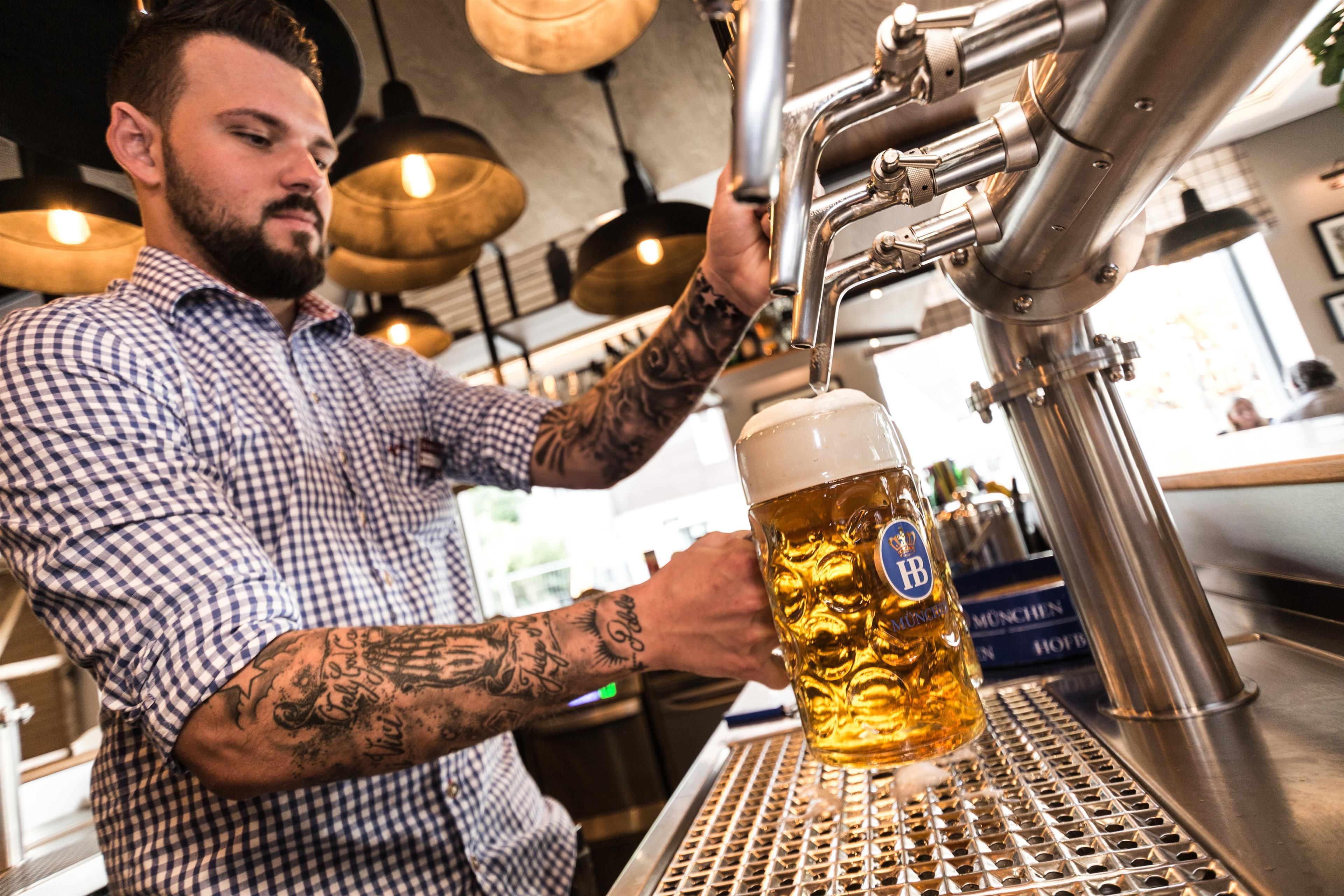 A person pouring beer into a mug at a bar with a metal grid counter and hanging lights in the background.