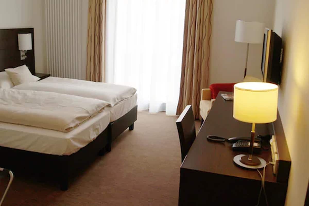 Quarto de hotel com duas camas, uma escrivaninha com abajur, uma televisão e uma janela com cortinas.