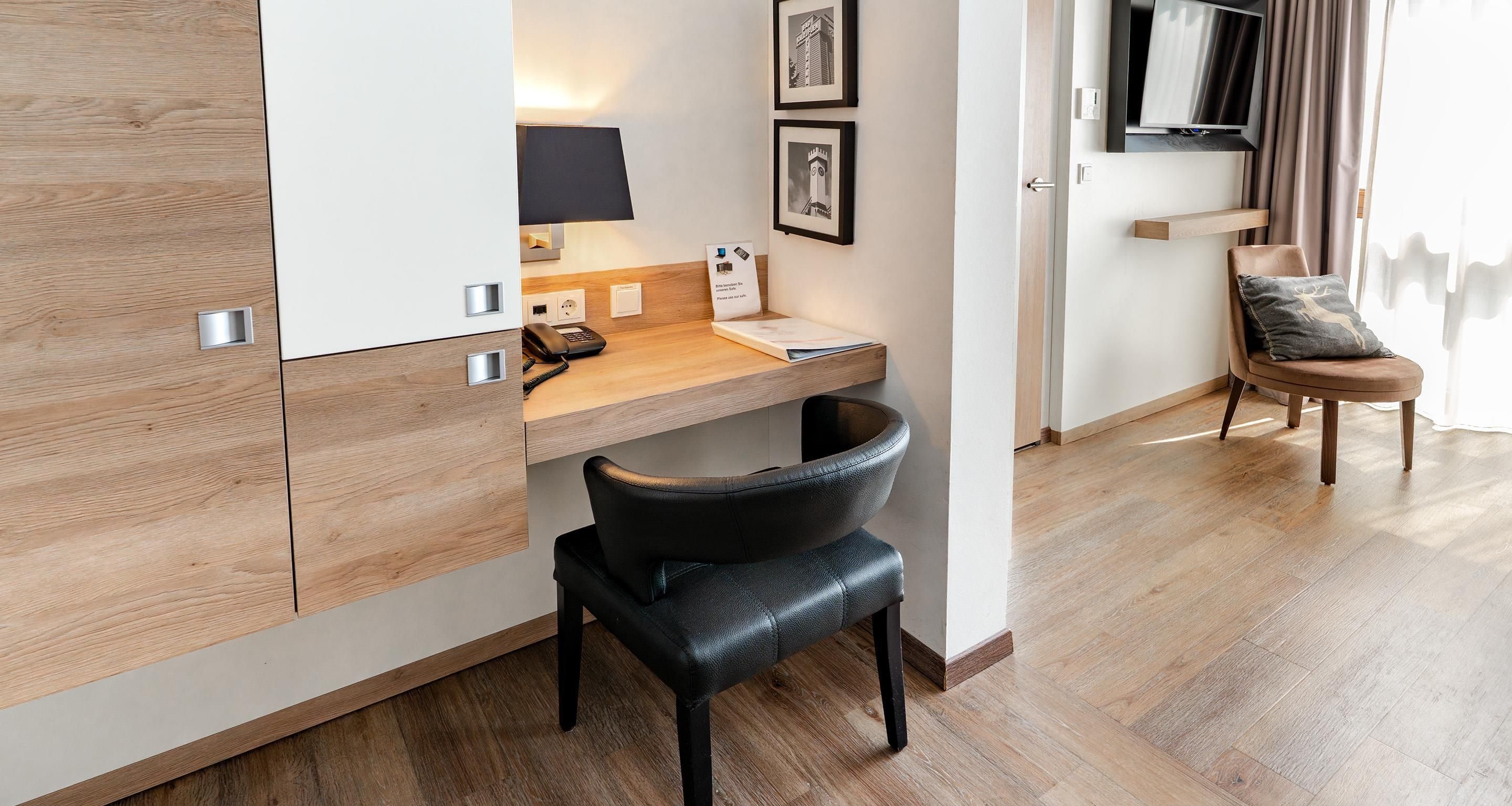 Ein modernes Hotelzimmer mit einem Holztisch, einem schwarzen Ledersessel, einem Flachbildfernseher und einem Bett mit einem Holz Kopfteil.