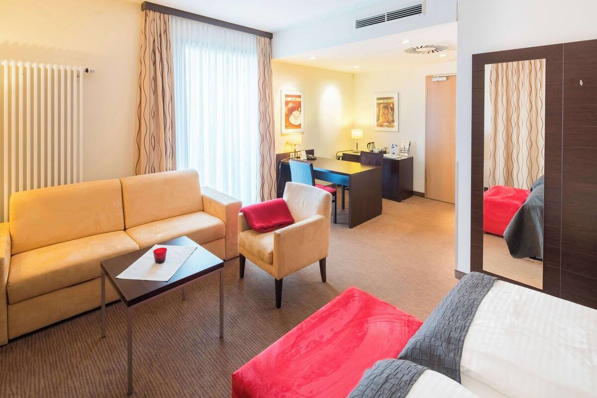 Une chambre d’hôtel avec un canapé, un fauteuil et un lit. On y trouve une petite table avec une tasse et une lampe. La chambre comprend également un coin repas avec une table et des chaises.
